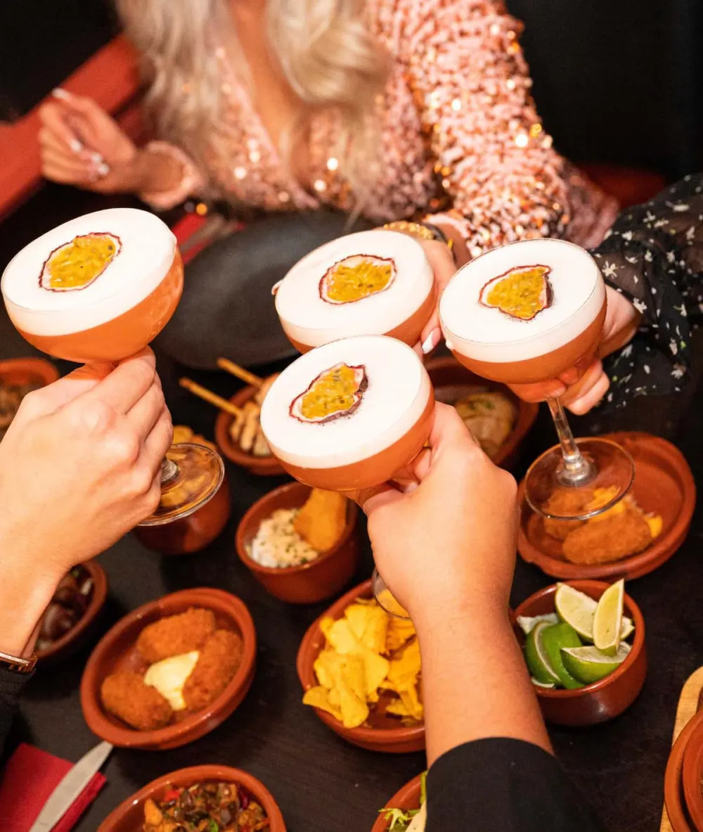 Vier gasten proosten met passievruchtcocktails boven een tafel vol tapas